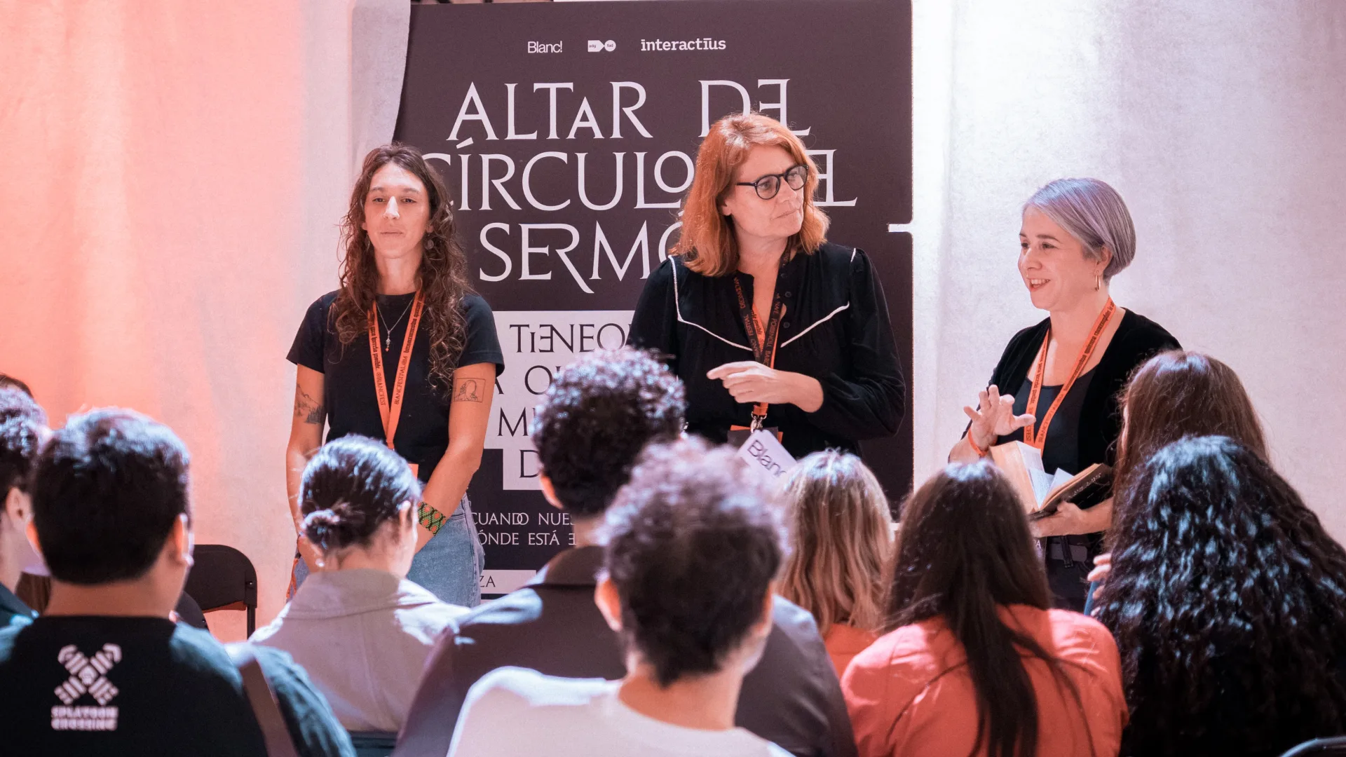 Tres dones dretes davant el plafó de l'Altar del Cercle del Sermó: la del centre parla gesticulant i la de la dreta sosté una llibreta. En primer terme, el públic assegut escolta d'esquena a la càmera.