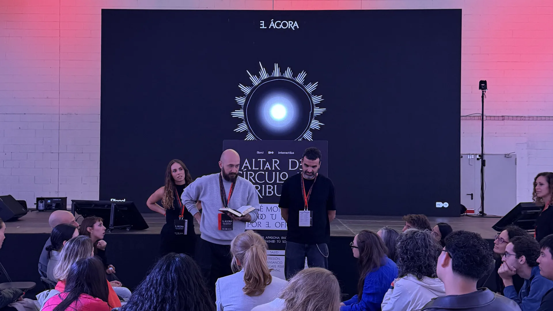 Tres persones dretes davant el plafó de l'Altar del Cercle del Tribut, una d'elles llegint d'un llibre. Al darrere, una pantalla gran amb el símbol lluminós d'El Dissenyor. El públic omple les cadires en primer terme.