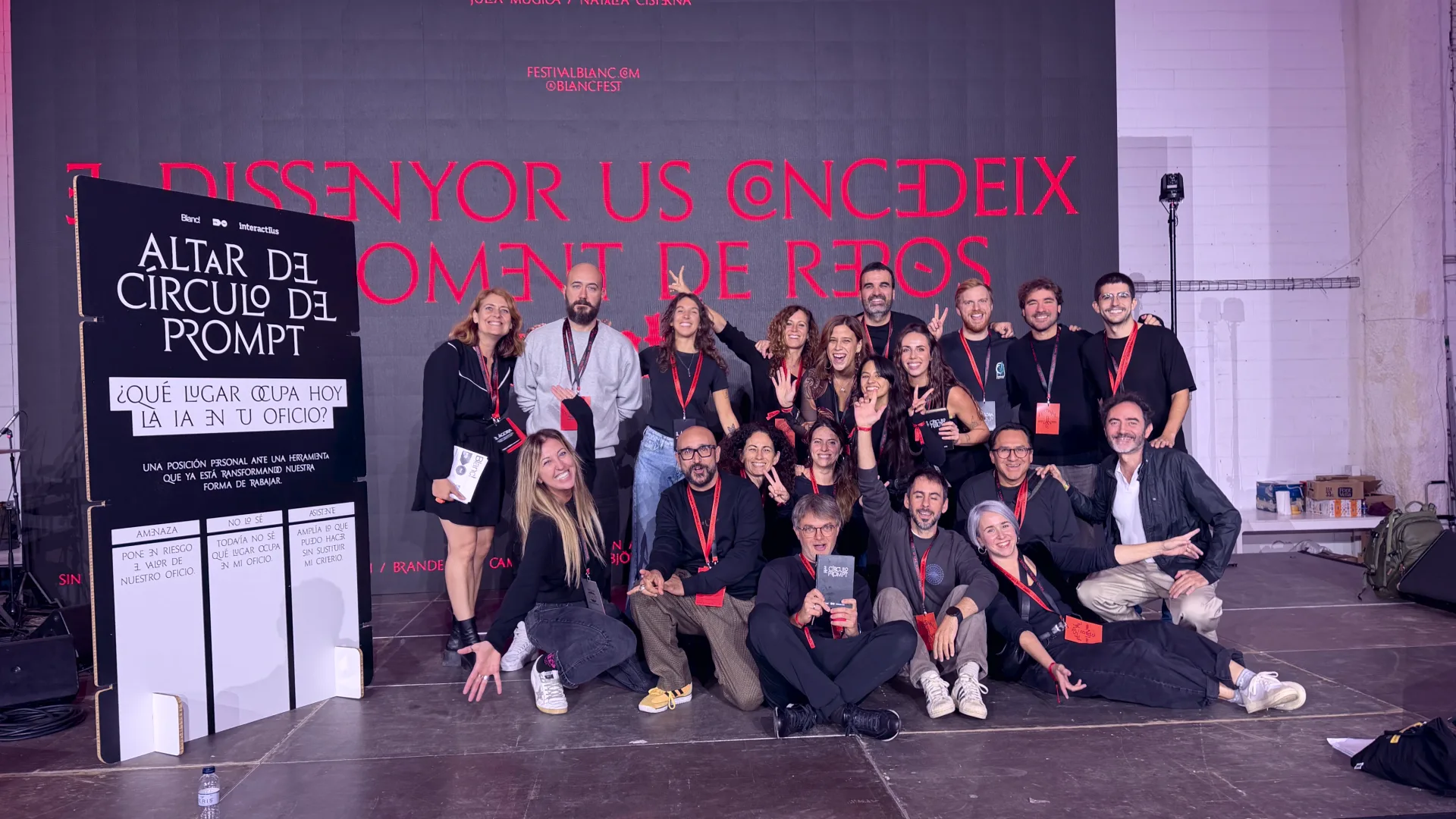Foto de grup de l'equip de l'Àgora, una vintena de persones posant entre rialles davant el plafó del Cercle del Prompt. Al fons, en lletres vermelles sobre la paret: «El Dissenyor us concedeix un moment de repòs».