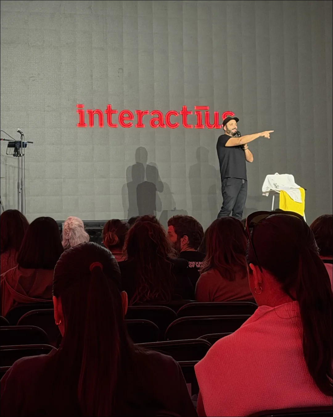 Una persona a l'escenari, micròfon a la mà, assenyalant el públic. Al darrere, el logotip d'Interactius projectat en vermell sobre la paret.